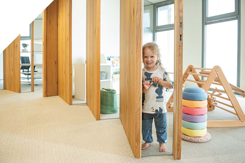 Ein kleines Kind steht barfuß und lächelnd in einem modernen, hellen Spielzimmer mit Holzbalken, bunt gestapelten Kissen und einem hölzernen Klettergerüst im Hintergrund.
