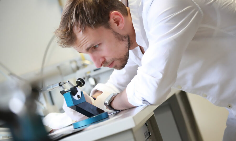 Ein Mann in einem weißen Hemd beugt sich über einen Labortisch und untersucht oder justiert mit konzentrierter Miene ein wissenschaftliches Gerät. Um ihn herum sind Laborgeräte und Drähte zu sehen.