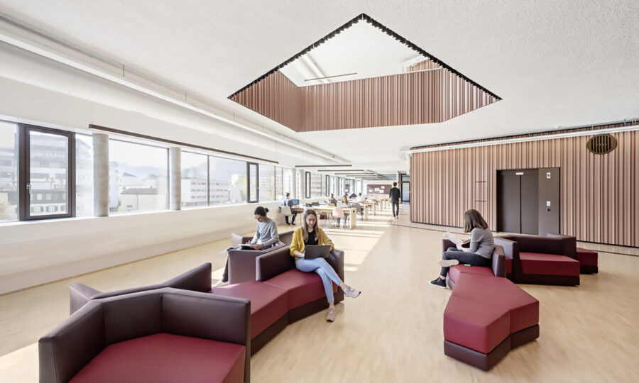 Eine moderne, helle Lounge mit großen Fenstern, geometrischen Sofas und Menschen, die sitzen oder Laptops benutzen. Der Raum verfügt über helle Holzböden, einen Deckenausschnitt und offene Bereiche zum Entspannen und Arbeiten.