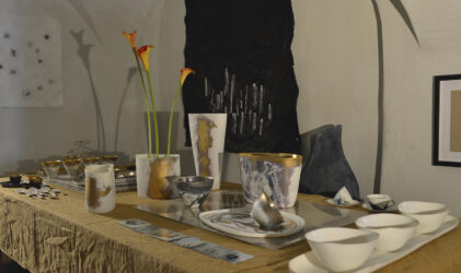 Auf einem mit einem hellbraunen Tuch bedeckten Ausstellungstisch stehen Keramikschalen, Vasen mit orangefarbenen Calla-Lilien, Tassen und Teller in Weiß-, Gold- und Grautönen, die wie in einer Kunstgalerie angeordnet sind.