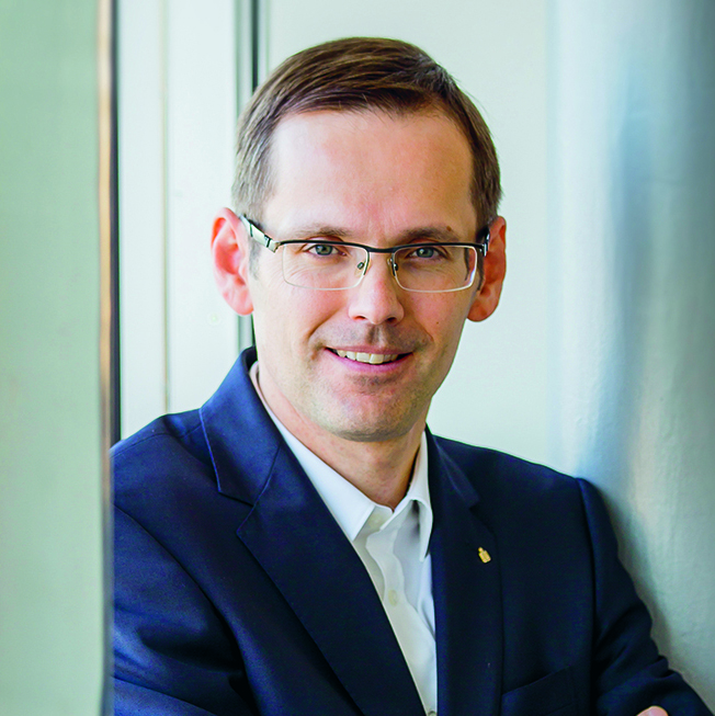 Ein Mann mit Brille, marineblauem Anzug und weißem Hemd steht in einem Haus, lehnt an einer Wand und lächelt in die Kamera.