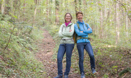 Zwei Frauen stehen mit verschränkten Armen auf einem Waldweg und lächeln in die Kamera. Sie tragen legere Outdoor-Kleidung und Rucksäcke. Bäume und Grünflächen umgeben sie, was auf eine Wanderung oder einen Naturspaziergang hindeutet.