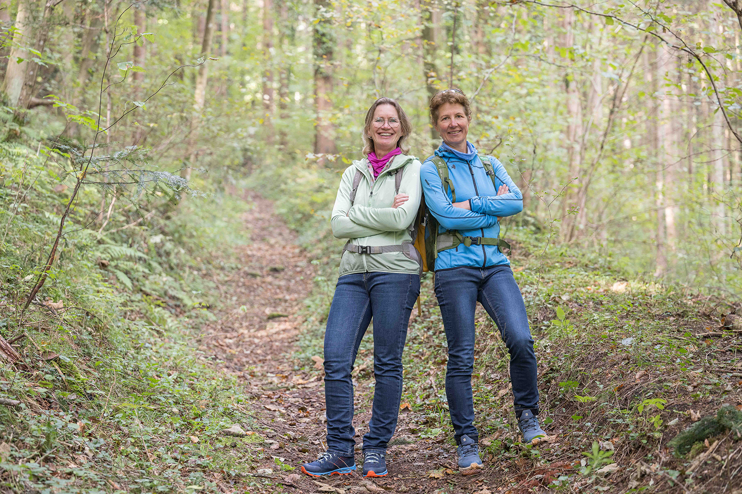 Zwei Frauen stehen mit verschränkten Armen auf einem Waldweg und lächeln in die Kamera. Sie tragen legere Outdoor-Kleidung und Rucksäcke. Bäume und Grünflächen umgeben sie, was auf eine Wanderung oder einen Naturspaziergang hindeutet.