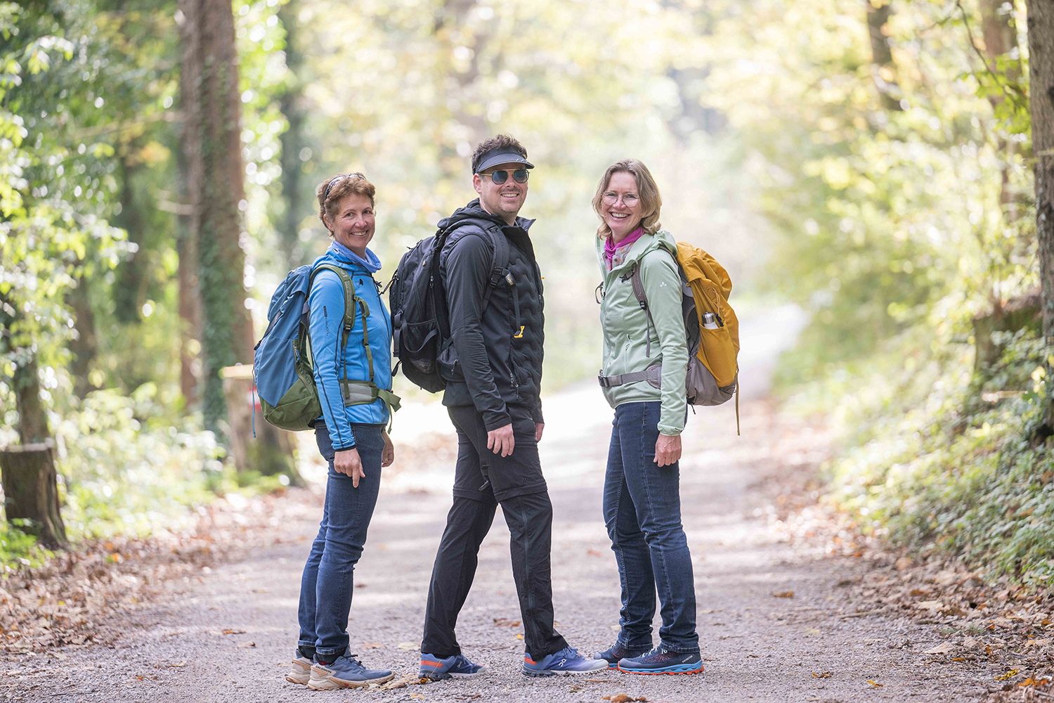 Drei Personen mit Rucksäcken stehen auf einem bewaldeten Weg und lächeln in die Kamera. Sie tragen legere Outdoor-Kleidung und sind von Bäumen umgeben, durch deren Blätter das Sonnenlicht fällt.