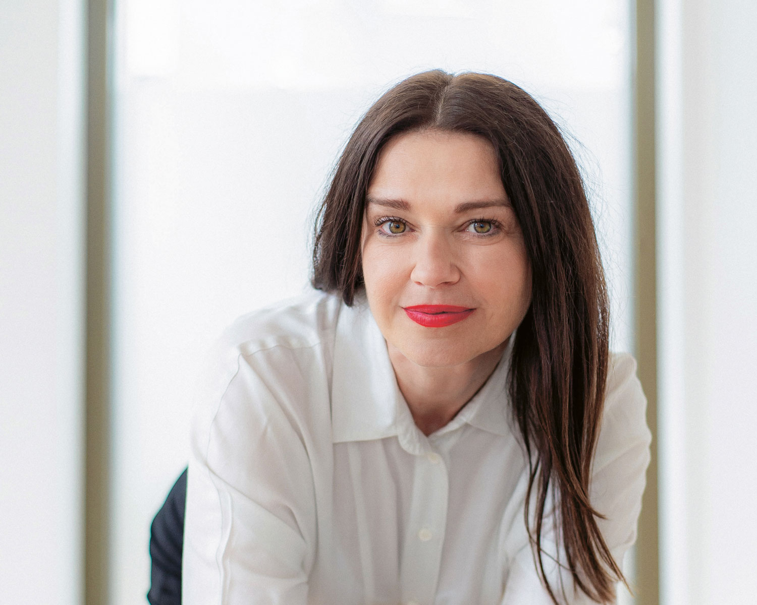 Eine Frau mit glattem dunklem Haar und rotem Lippenstift, die ein weißes Button-up-Hemd trägt, lächelt in einem hell erleuchteten Raum mit unscharfem Hintergrund sanft in die Kamera.