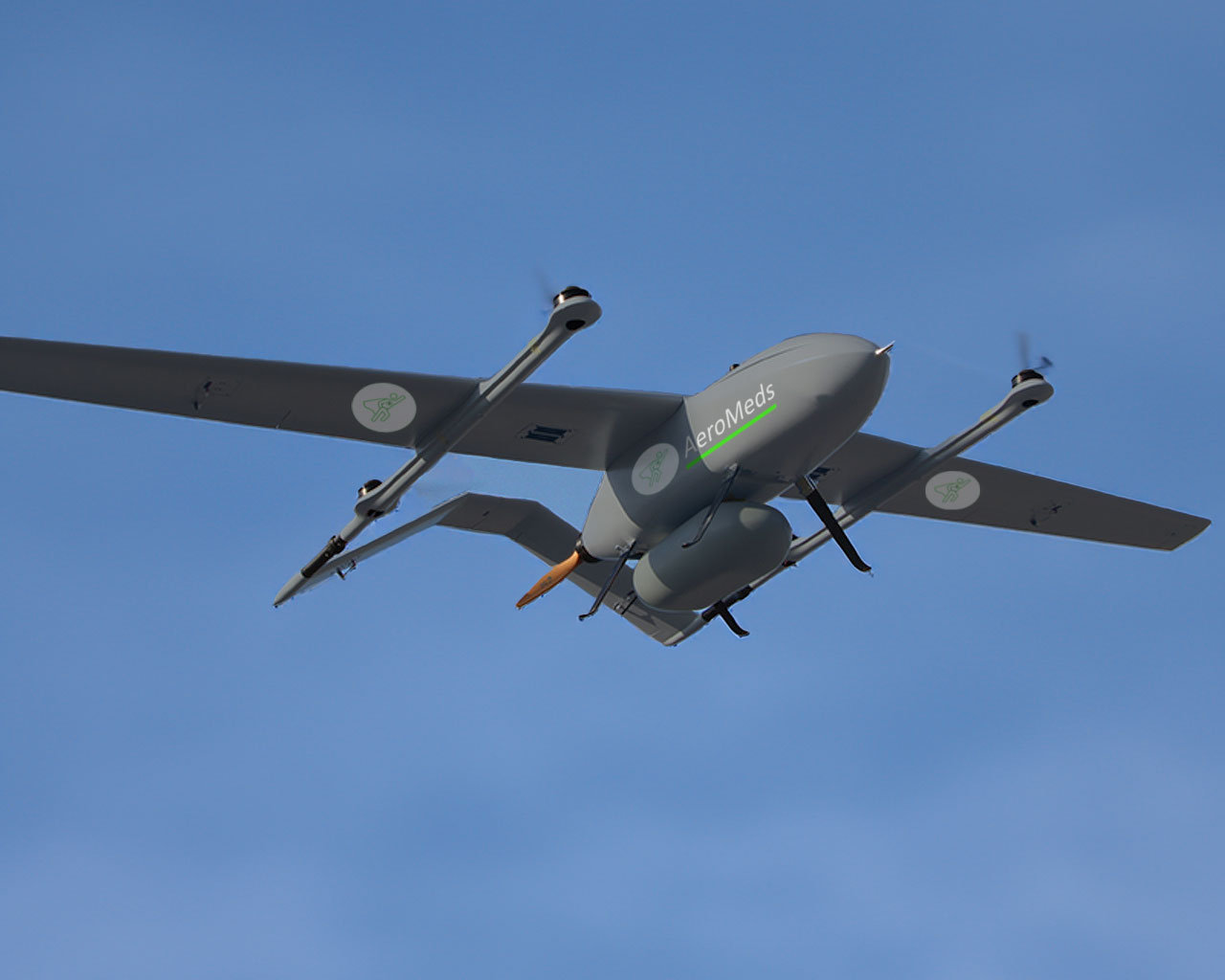 Eine graue Drohne mit dem Namen "AeroMeds" und grünen kreisförmigen Logos auf dem Körper und den Flügeln fliegt gegen einen klaren blauen Himmel. Die Drohne hat zwei Propeller und scheint eine Nutzlast unter sich zu tragen.