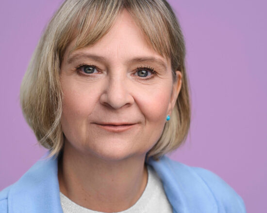 Eine Frau mittleren Alters mit kurzem blondem Haar lächelt sanft in die Kamera. Sie trägt eine hellblaue Jacke und ein weißes Oberteil, dahinter ein lavendelfarbener Hintergrund.