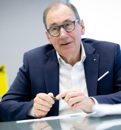 Ein Mann mittleren Alters, der eine Brille und einen marineblauen Anzug trägt, sitzt an einem Schreibtisch mit Papieren, hält einen Stift in der Hand und scheint während einer Besprechung zu sprechen. Ein Glas Wasser und ein gelber Gegenstand stehen auf dem Tisch.