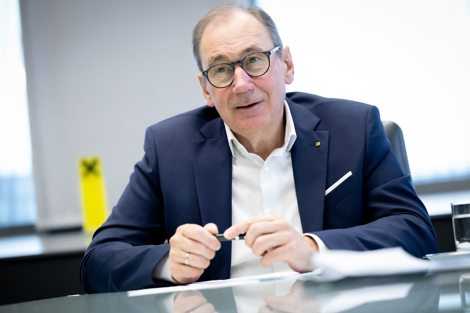 Ein Mann mittleren Alters, der eine Brille und einen marineblauen Anzug trägt, sitzt an einem Schreibtisch mit Papieren, hält einen Stift in der Hand und scheint während einer Besprechung zu sprechen. Ein Glas Wasser und ein gelber Gegenstand stehen auf dem Tisch.