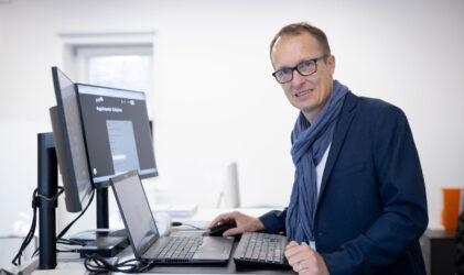 Ein Mann, der eine Brille, einen blauen Blazer und einen Schal trägt, steht an einem Schreibtisch mit zwei Monitoren und einem Laptop und lächelt in einem hellen, modernen Büro in die Kamera.