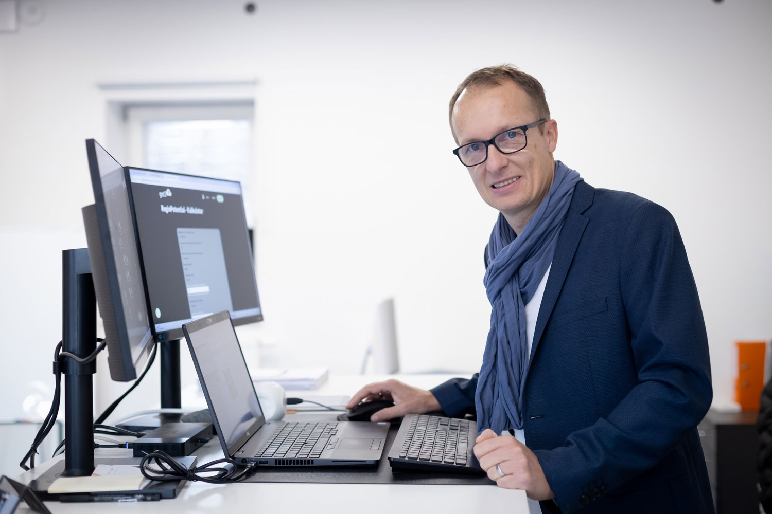 Ein Mann, der eine Brille, einen blauen Blazer und einen Schal trägt, steht an einem Schreibtisch mit zwei Monitoren und einem Laptop und lächelt in einem hellen, modernen Büro in die Kamera.