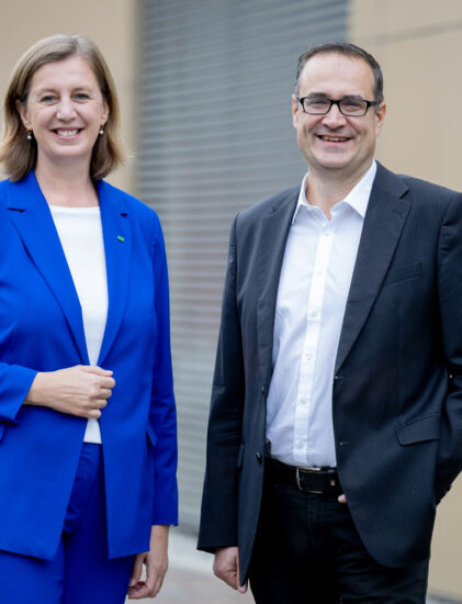 Zwei Personen stehen im Freien und lächeln in die Kamera. Die Frau auf der linken Seite trägt einen hellblauen Anzug, der Mann auf der rechten Seite eine Brille, ein weißes Hemd und einen schwarzen Blazer. Im Hintergrund sind eine beigefarbene Wand und Metallfensterläden zu sehen.
