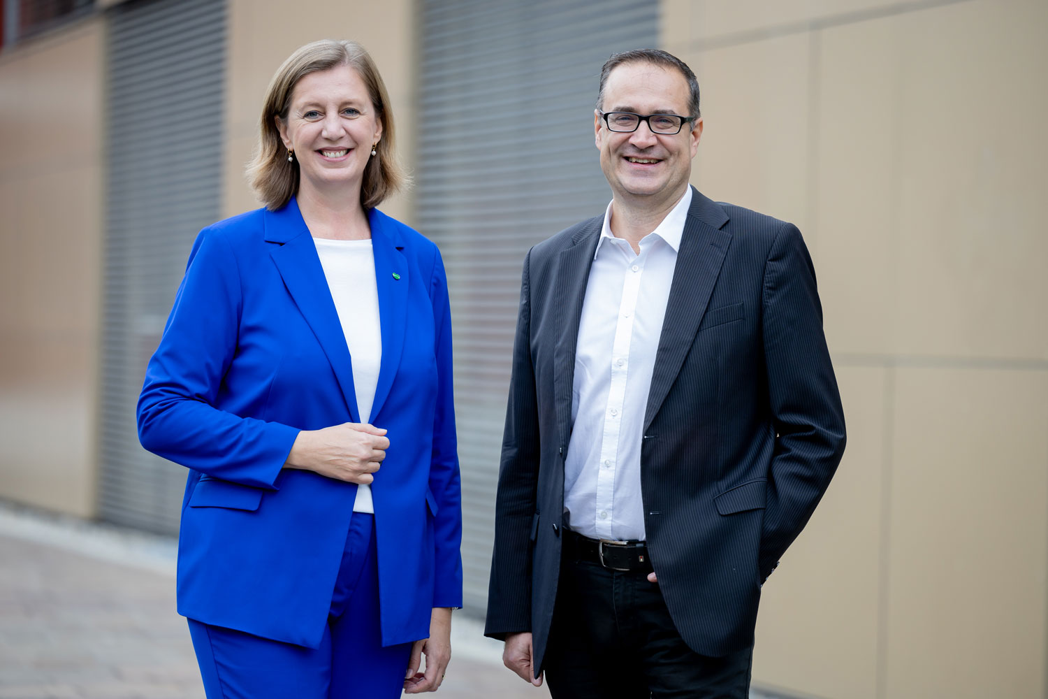 Zwei Personen stehen im Freien und lächeln in die Kamera. Die Frau auf der linken Seite trägt einen hellblauen Anzug, der Mann auf der rechten Seite eine Brille, ein weißes Hemd und einen schwarzen Blazer. Im Hintergrund sind eine beigefarbene Wand und Metallfensterläden zu sehen.