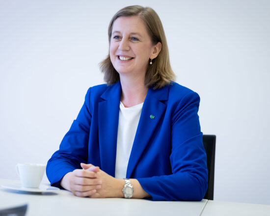 Eine Frau mit schulterlangem hellbraunem Haar sitzt lächelnd an einem Tisch. Sie trägt einen hellblauen Blazer über einem weißen Oberteil, eine Uhr und Ohrringe. Auf dem Tisch vor ihr stehen eine weiße Tasse und eine Untertasse.
