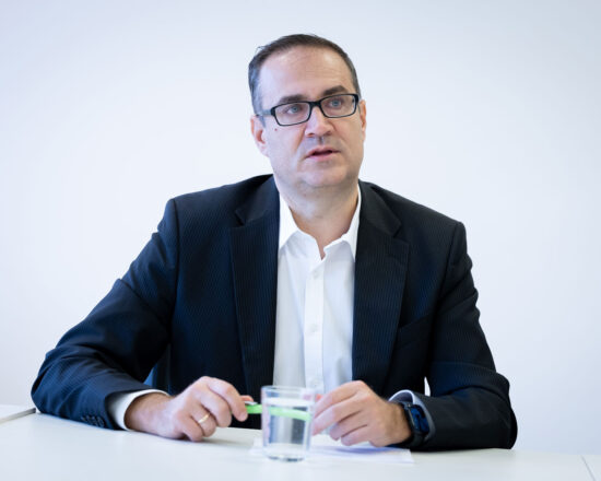 Ein Mann im dunklen Anzug und mit Brille sitzt an einem weißen Tisch, hält einen Stift und ein Glas Wasser in der Hand und blickt leicht von der Kamera weg vor einem schlichten hellen Hintergrund.