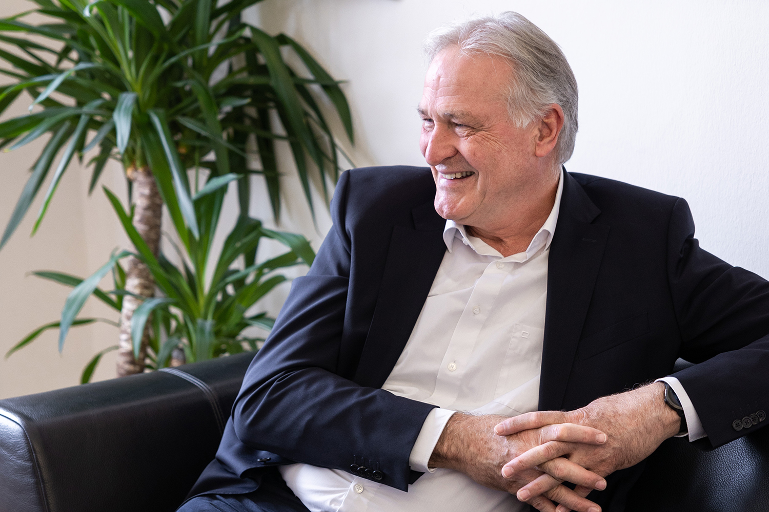 Ein lächelnder älterer Mann in einem dunklen Anzug sitzt in einem hellen Raum auf einer schwarzen Ledercouch mit gefalteten Händen neben einer hohen Grünpflanze.