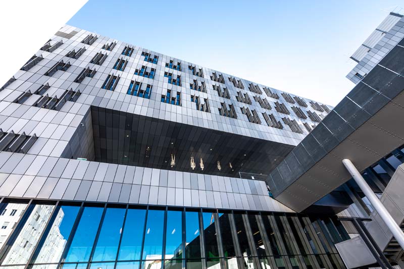Modernes mehrstöckiges Gebäude mit einer Glasfassade und geometrischen Metallpaneelen, von unten gesehen vor einem klaren blauen Himmel. In den Fenstern sind Spiegelungen sichtbar.