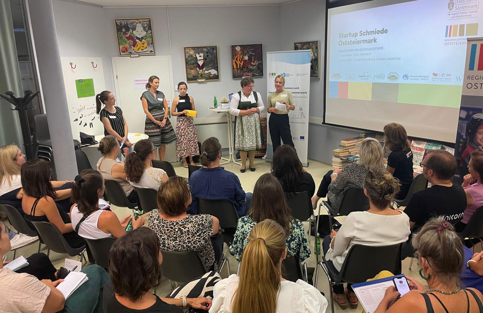 Eine Gruppe von Frauen steht vorne in einem Klassenzimmer und hält einen Vortrag vor einem Publikum. Auf einer Leinwand ist "Startup Schmiede Oststeiermark" zu sehen, und die Zuhörer sitzen, hören zu und machen sich Notizen. An den Wänden hängen Kunstwerke.