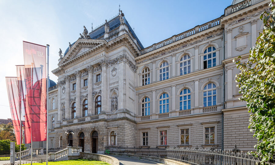 Ein prächtiges historisches Gebäude mit verschnörkelten architektonischen Details und Bogenfenstern unter strahlend blauem Himmel. Vor dem Gebäude sind drei rot-weiße Fahnen an Masten angebracht, und ein geschwungener Weg führt zum Eingang.