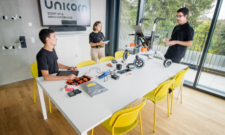 Drei Personen arbeiten in einem hellen Büro mit der Aufschrift "Unicorn Start-Up & Innovation Hub" und diskutieren über einen fahrbaren Roboter auf einem Tisch mit Elektronik, Werkzeugen und Laptops, während große Fenster den Blick ins Grüne freigeben.