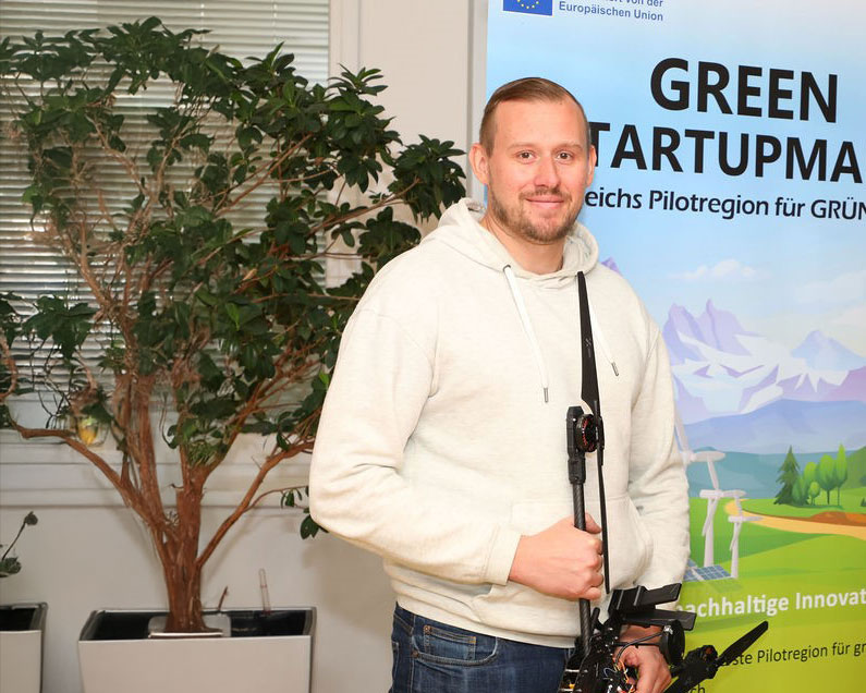 Ein Mann in einem hellen Kapuzenpulli steht in einem Haus und hält eine Kameraausrüstung. Hinter ihm steht eine große Topfpflanze und ein buntes Banner mit der Aufschrift "GREEN STARTUPMA" und Bildern von Windturbinen und Solarzellen.