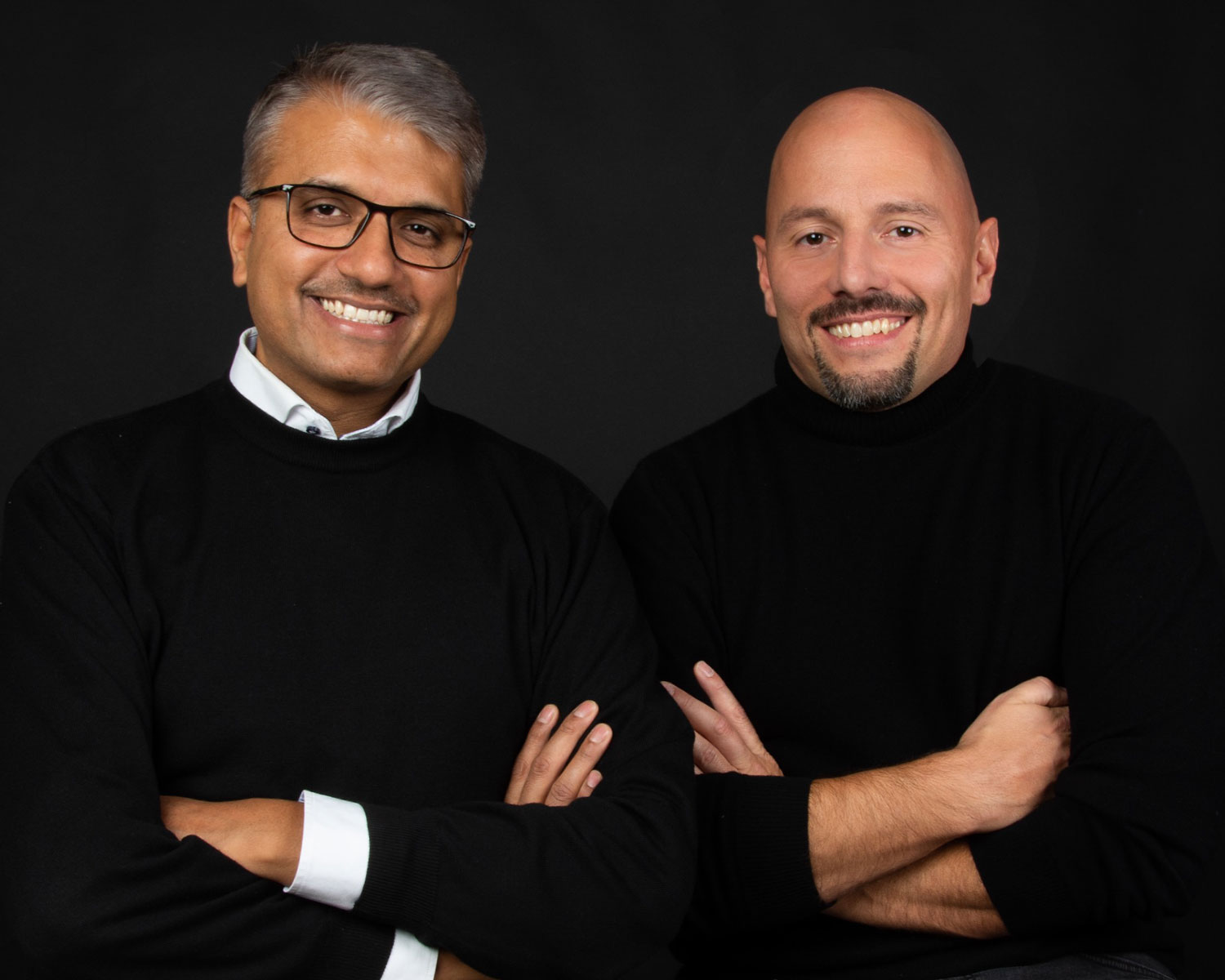 Zwei Männer lächeln und posieren zusammen vor einem schwarzen Hintergrund. Einer hat eine Brille und graues Haar, der andere eine Glatze. Beide tragen schwarze Pullover und haben die Arme verschränkt.