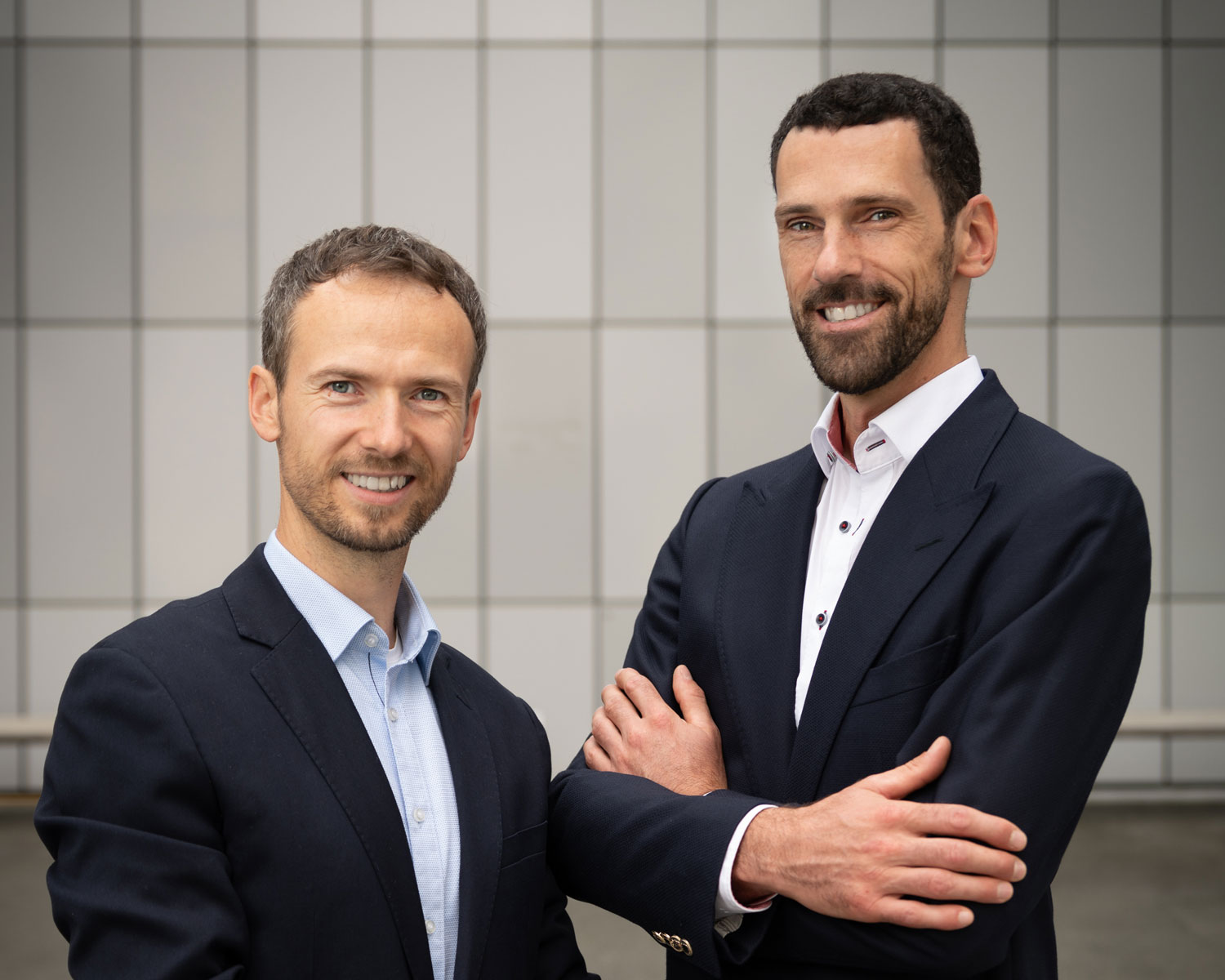 Zwei Männer in Geschäftsanzügen stehen Seite an Seite und lächeln selbstbewusst in die Kamera. Der eine hat die Arme verschränkt, der andere hat die Hände an den Seiten. Sie stehen vor einem modernen, gekachelten Wandhintergrund.
