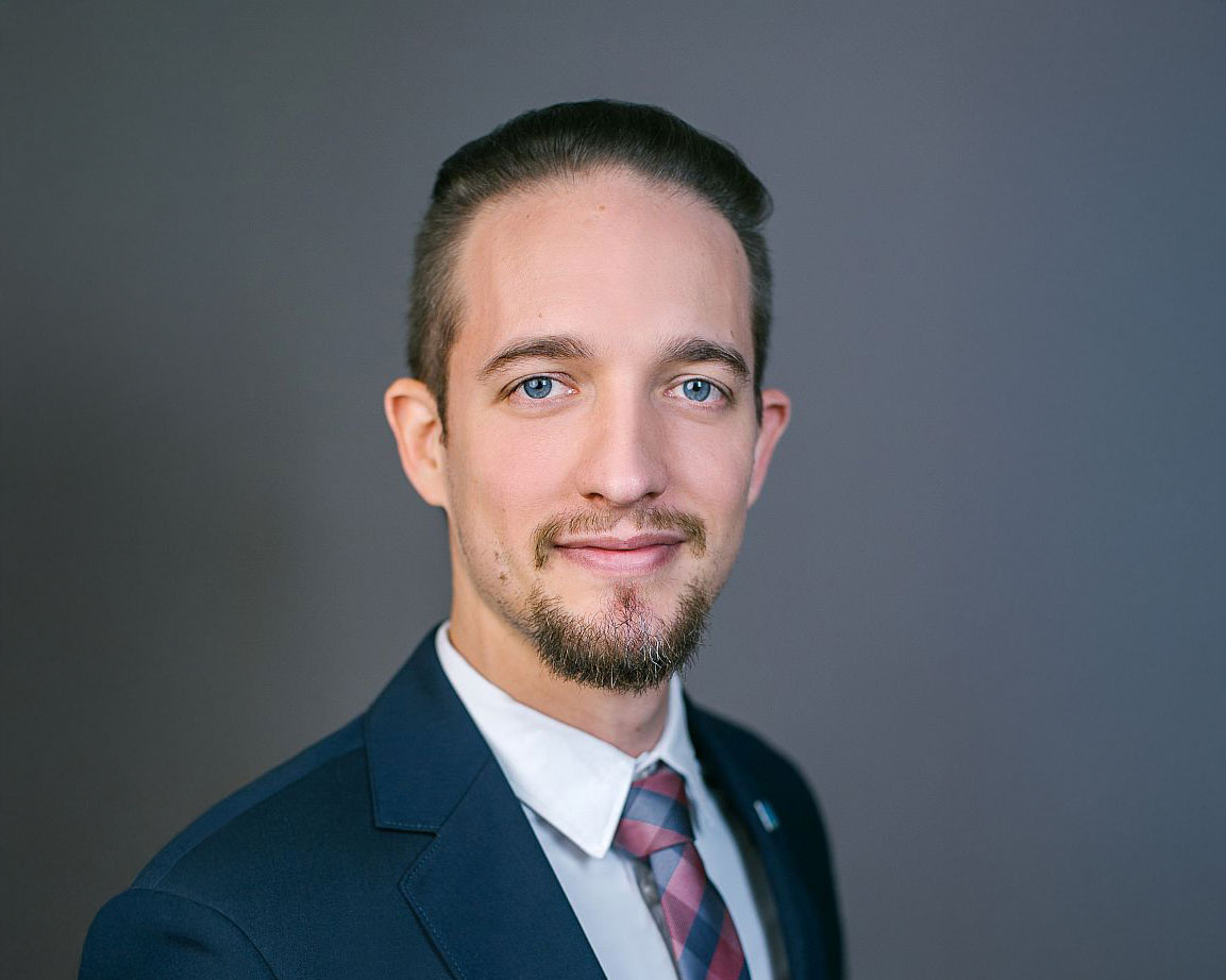 Ein Mann in Anzug und Krawatte mit gepflegtem Haar und kurzem Bart, der leicht lächelt, posiert vor einem schlichten grauen Hintergrund.