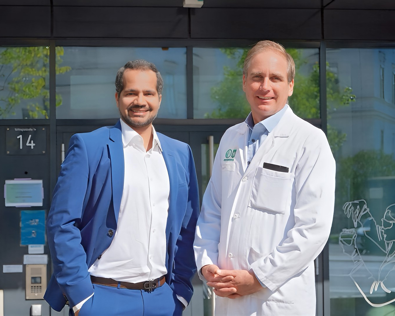 Zwei Männer stehen Seite an Seite vor einem Gebäude. Der eine trägt einen blauen Anzug und ein weißes Hemd, der andere einen weißen Arztkittel. Beide lächeln und schauen in die Kamera. Im Hintergrund sind Glastüren und Grünflächen zu sehen.