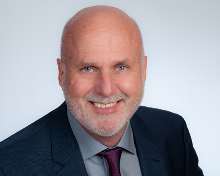 Ein lächelnder Mann mittleren Alters mit Glatze und kurzem grauem Bart, der einen dunklen Anzug, ein graues Hemd und eine kastanienbraune Krawatte trägt, posiert vor einem schlichten hellen Hintergrund.