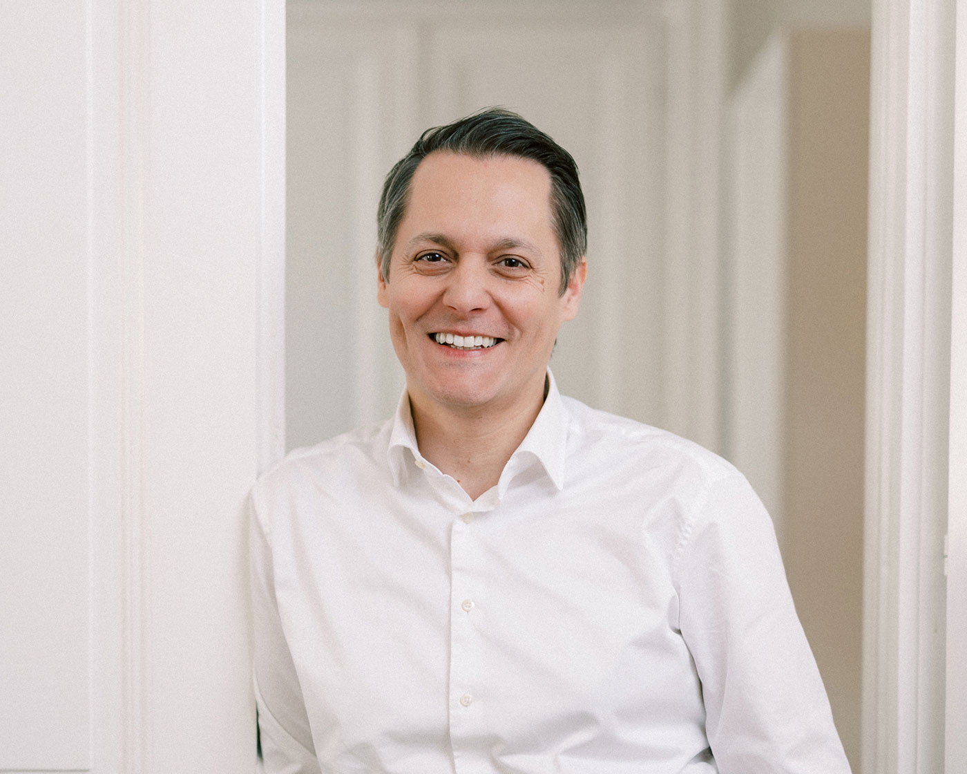 Ein Mann mit kurzen dunklen Haaren und weißem Hemd steht in einem schwach beleuchteten Raum neben einer weißen Wand und lächelt freundlich.