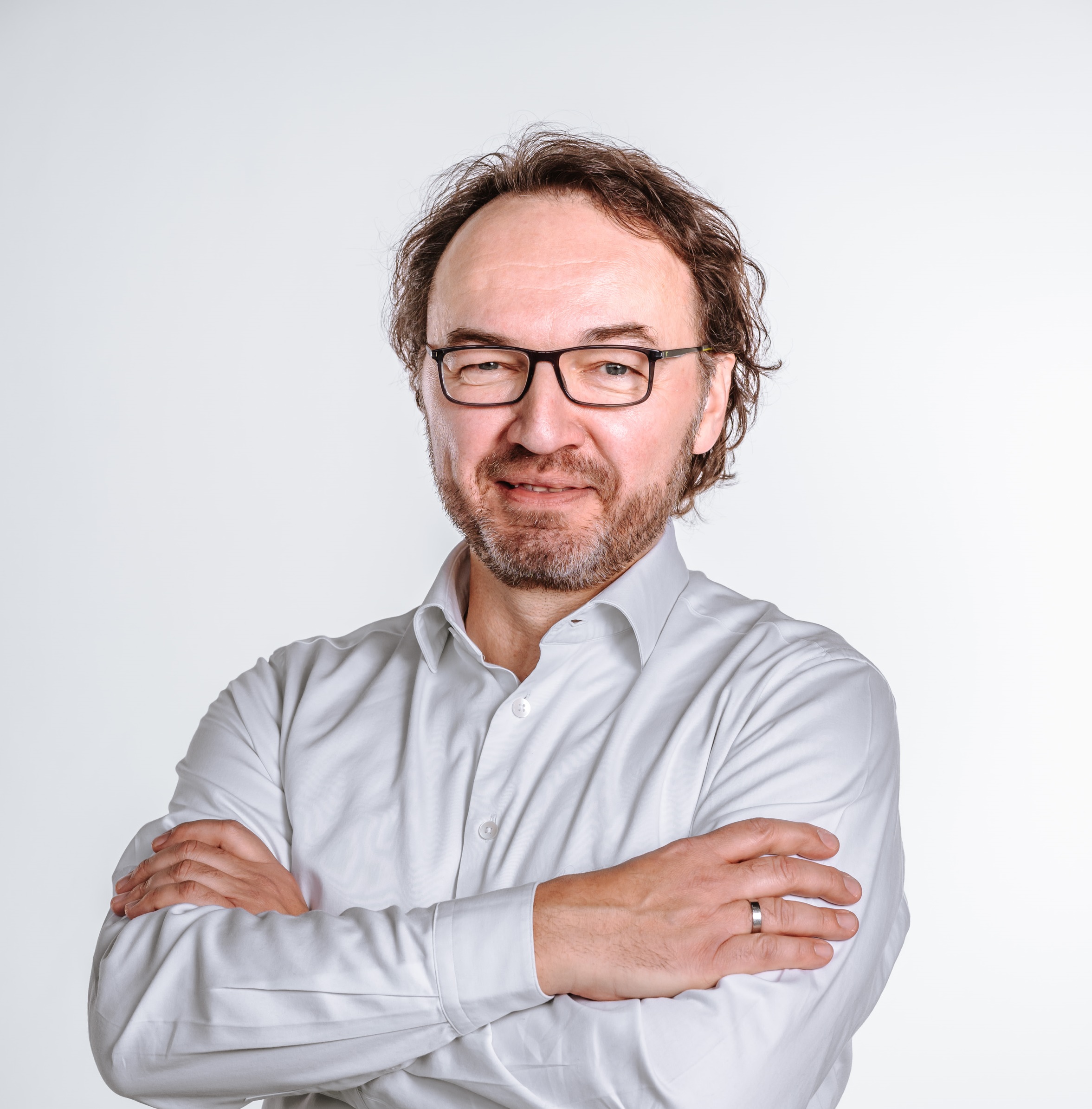 Ein Mann mittleren Alters mit Brille und Bart steht vor einem schlichten weißen Hintergrund und lächelt leicht mit verschränkten Armen. Er trägt ein weißes Button-up-Hemd.
