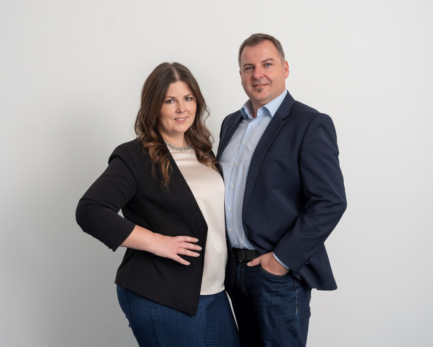 Eine Frau und ein Mann stehen Seite an Seite vor einem schlichten weißen Hintergrund, beide tragen dunkle Blazer und Jeans. Die Frau hat langes braunes Haar und ihre Hand in die Hüfte gestemmt, während der Mann lächelt und eine Hand in der Tasche hat.