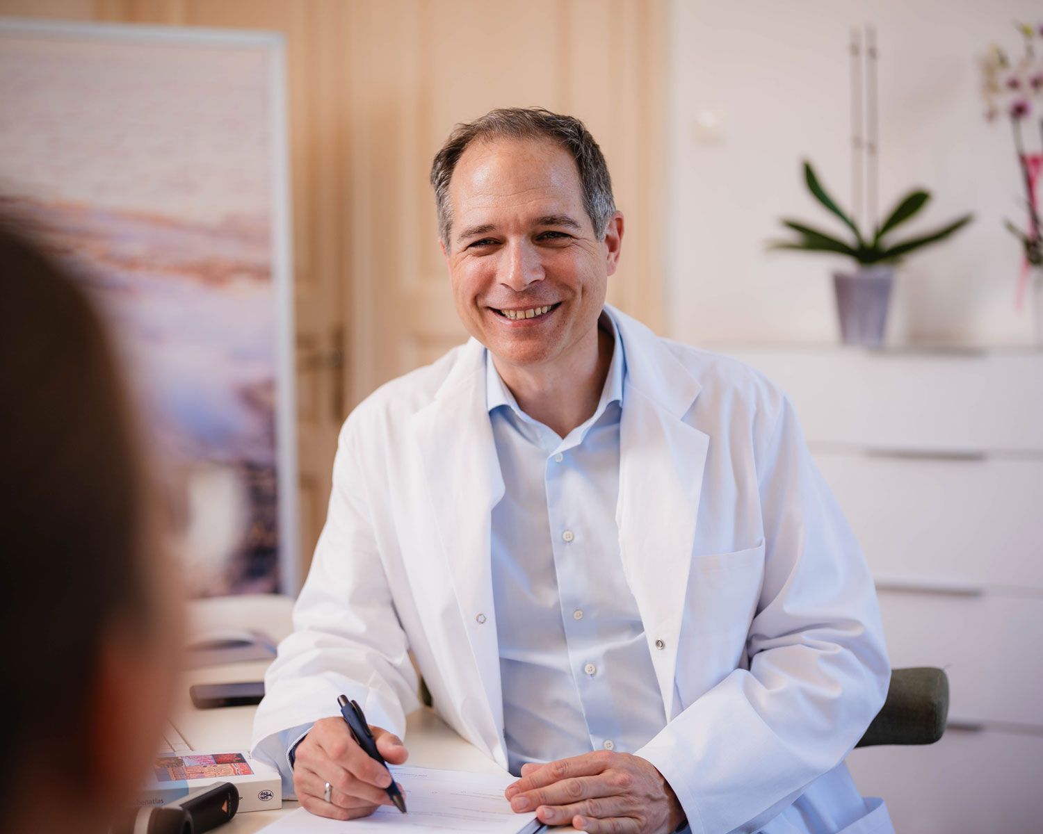 Ein lächelnder Arzt im weißen Kittel sitzt am Schreibtisch, hält Stift und Papier in der Hand und unterhält sich mit einem Patienten in einem hellen, modernen Büro mit Pflanzen und sanfter Beleuchtung.