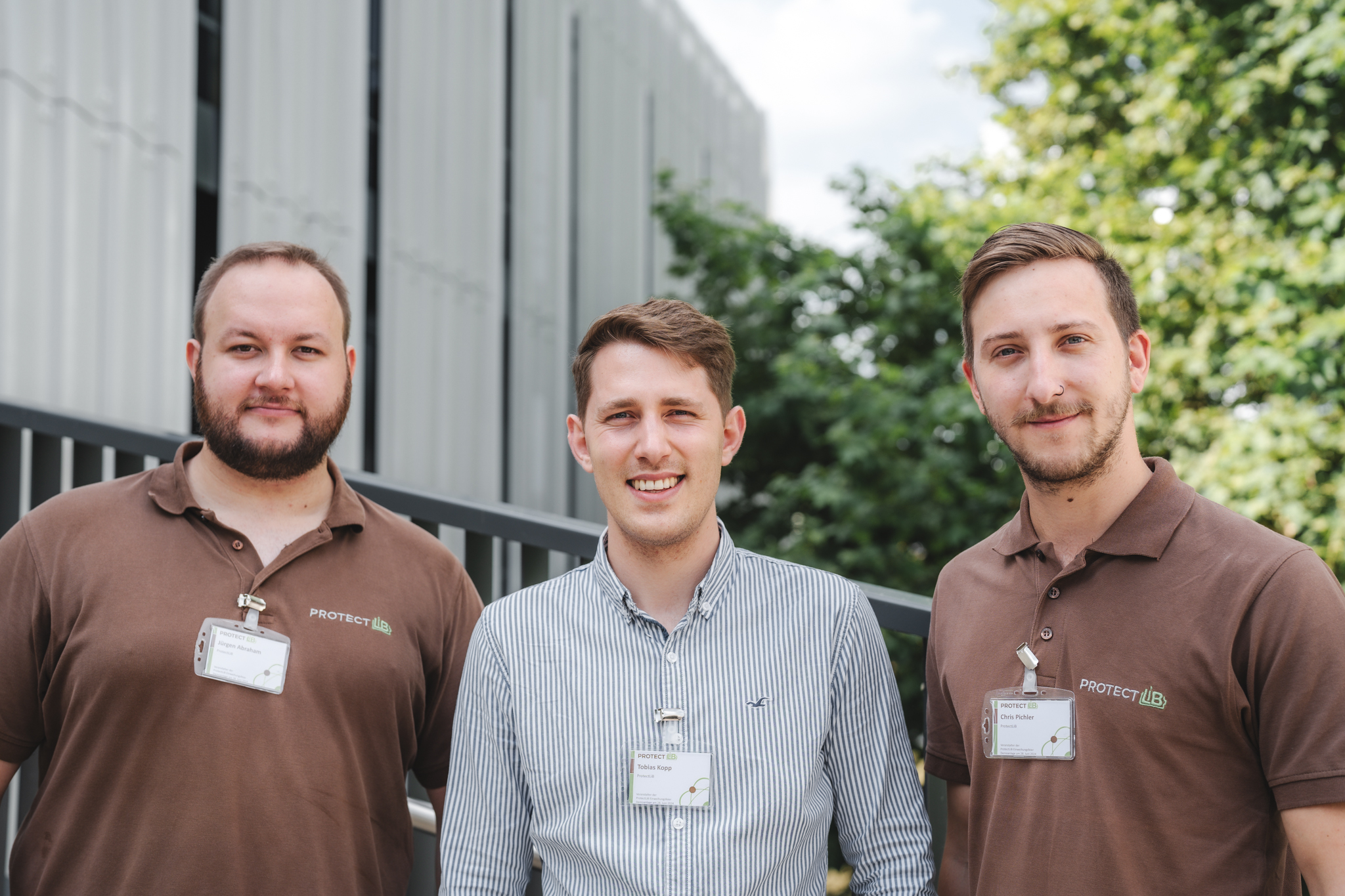 Drei Männer stehen im Freien, zwei in braunen Poloshirts mit Namensschildern und einer in einem gestreiften Button-up-Hemd. Hinter ihnen sind ein modernes Gebäude und grüne Bäume zu sehen. Alle lächeln in die Kamera.