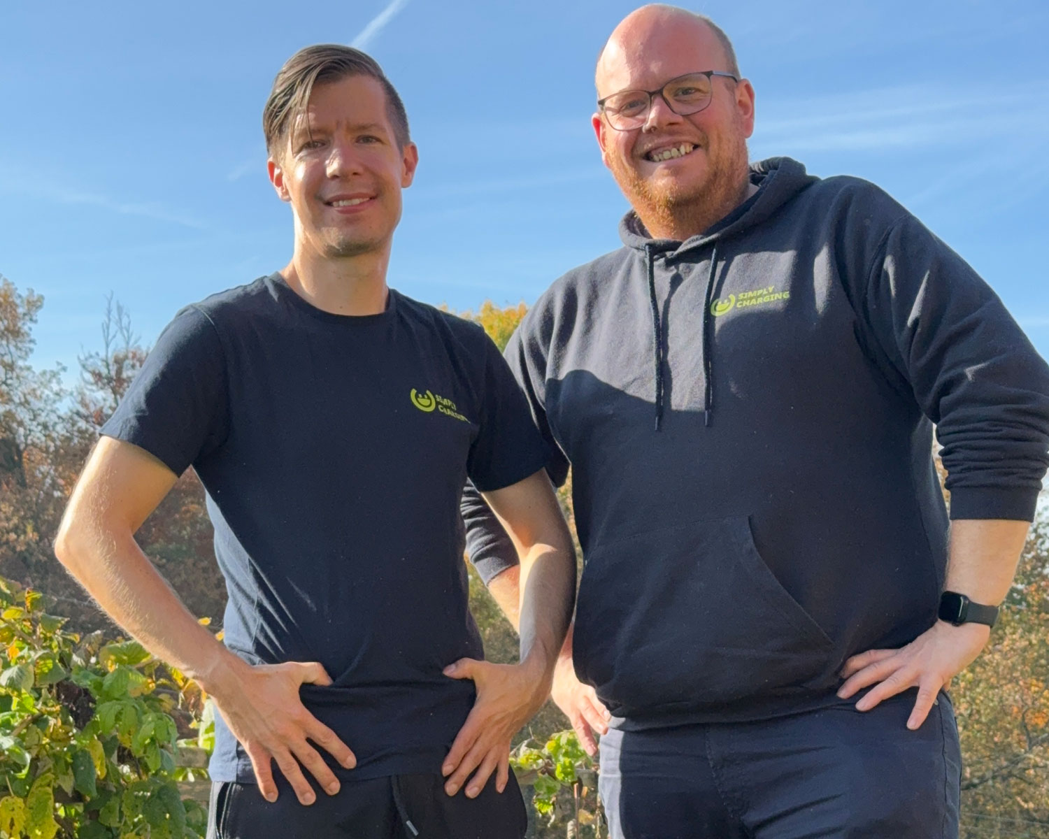 Zwei Männer stehen an einem sonnigen Tag im Freien und lächeln in die Kamera. Beide sind leger in dunkle Hemden gekleidet und haben die Hände in die Hüften gestemmt. Im Hintergrund sind Bäume mit Herbstlaub zu sehen.