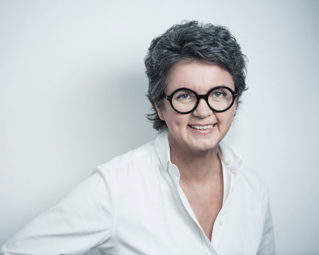 Eine lächelnde Person mit kurzen, lockigen grauen Haaren und einer runden schwarzen Brille, die ein weißes Button-up-Hemd trägt, steht vor einem schlichten hellen Hintergrund.
