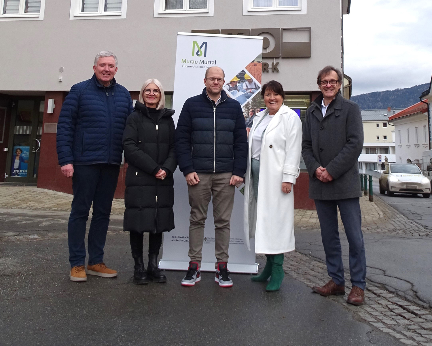 Fünf Personen stehen im Freien vor einem "Murau Murtal"-Transparent auf einer Stadtstraße, sie tragen Winterkleidung und lächeln in die Kamera. Im Hintergrund sind Gebäude und ein geparktes Auto zu sehen.