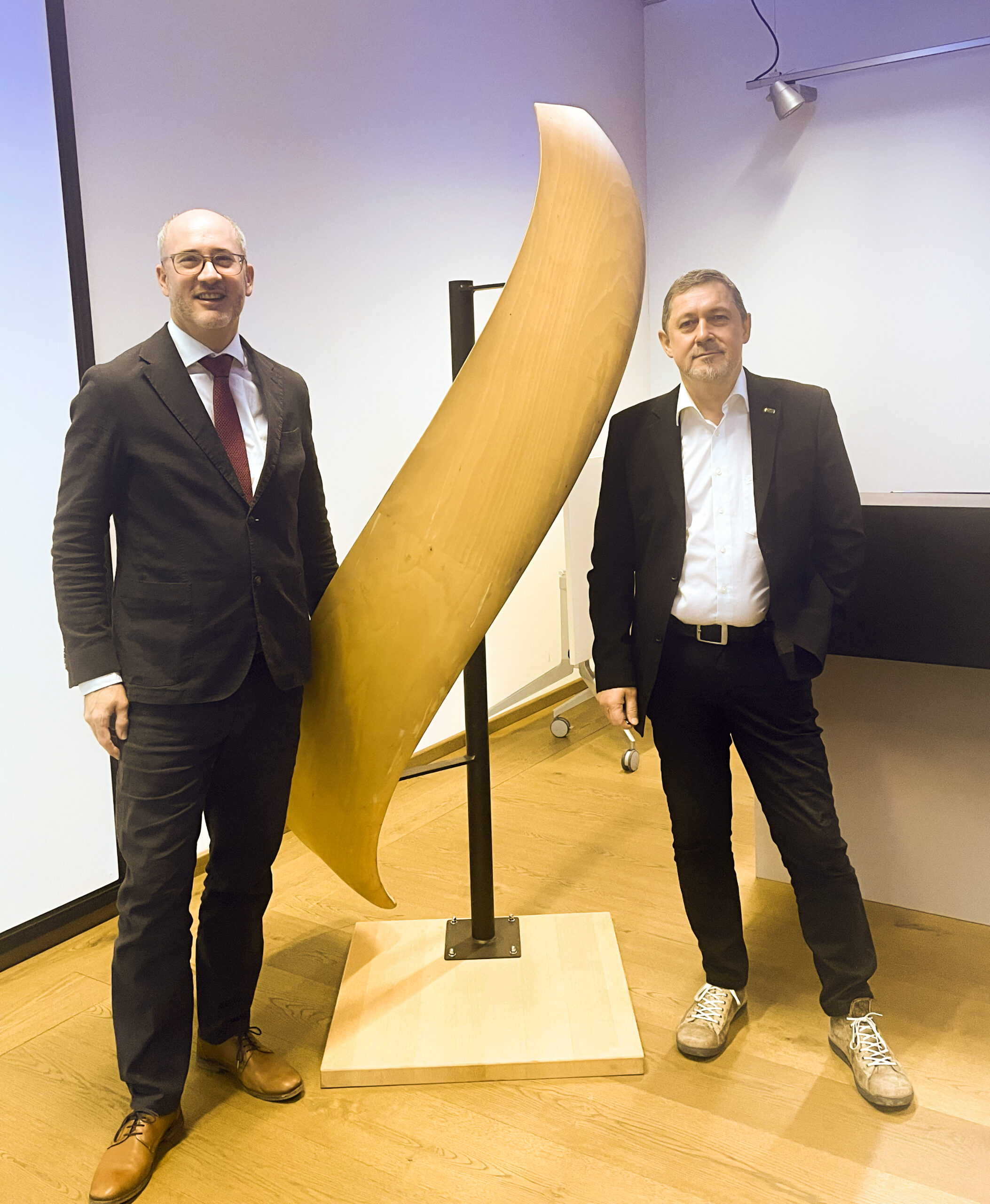 Zwei Männer in Anzügen stehen in einem Innenraum auf beiden Seiten einer großen, geschwungenen Holzskulptur auf einem Sockel. Die Umgebung scheint ein formeller oder akademischer Raum mit Holzboden und weißen Wänden zu sein.