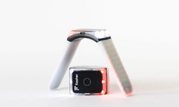 Ein tragbares Gerät mit einem flexiblen, weißen Band, das mit roten und weißen Lichtern leuchtet, ist in einem Bogen über einem kleinen schwarzen, rechteckigen Gerät mit der Aufschrift "Flesher" vor einem schlichten weißen Hintergrund gebogen.