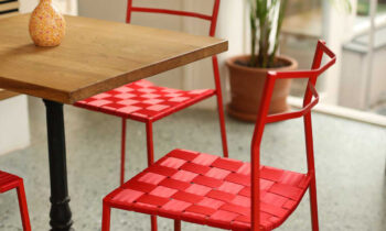 Ein Holztisch mit einer Blumenvase ist von roten Metallstühlen mit geflochtenen Sitzen umgeben, die auf einem gesprenkelten Boden neben einer Topfpflanze und großen sonnigen Fenstern stehen.