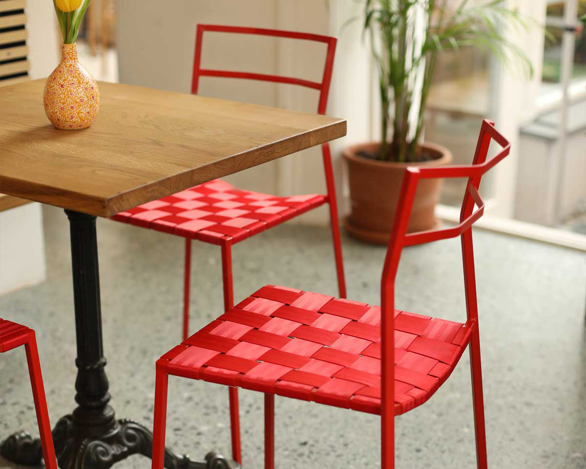 Ein Holztisch mit einer Blumenvase ist von roten Metallstühlen mit geflochtenen Sitzen umgeben, die auf einem gesprenkelten Boden neben einer Topfpflanze und großen sonnigen Fenstern stehen.