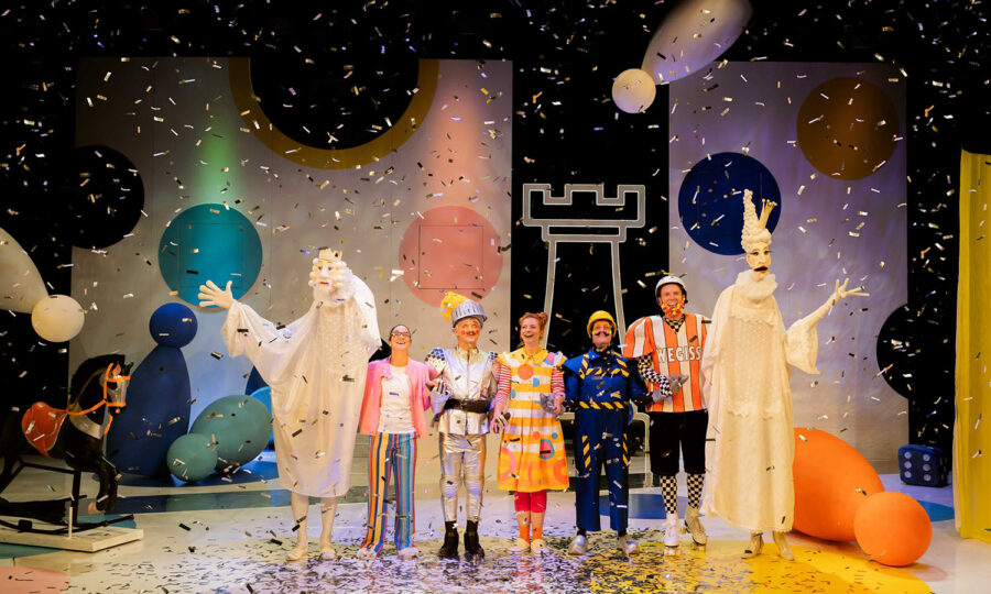 Eine Gruppe von Darstellern in farbenfrohen, skurrilen Kostümen steht auf einer verspielten Bühne mit überdimensionalen Spielfiguren, Konfetti fällt um sie herum, während sie unter hellen Lichtern posieren und lächeln.