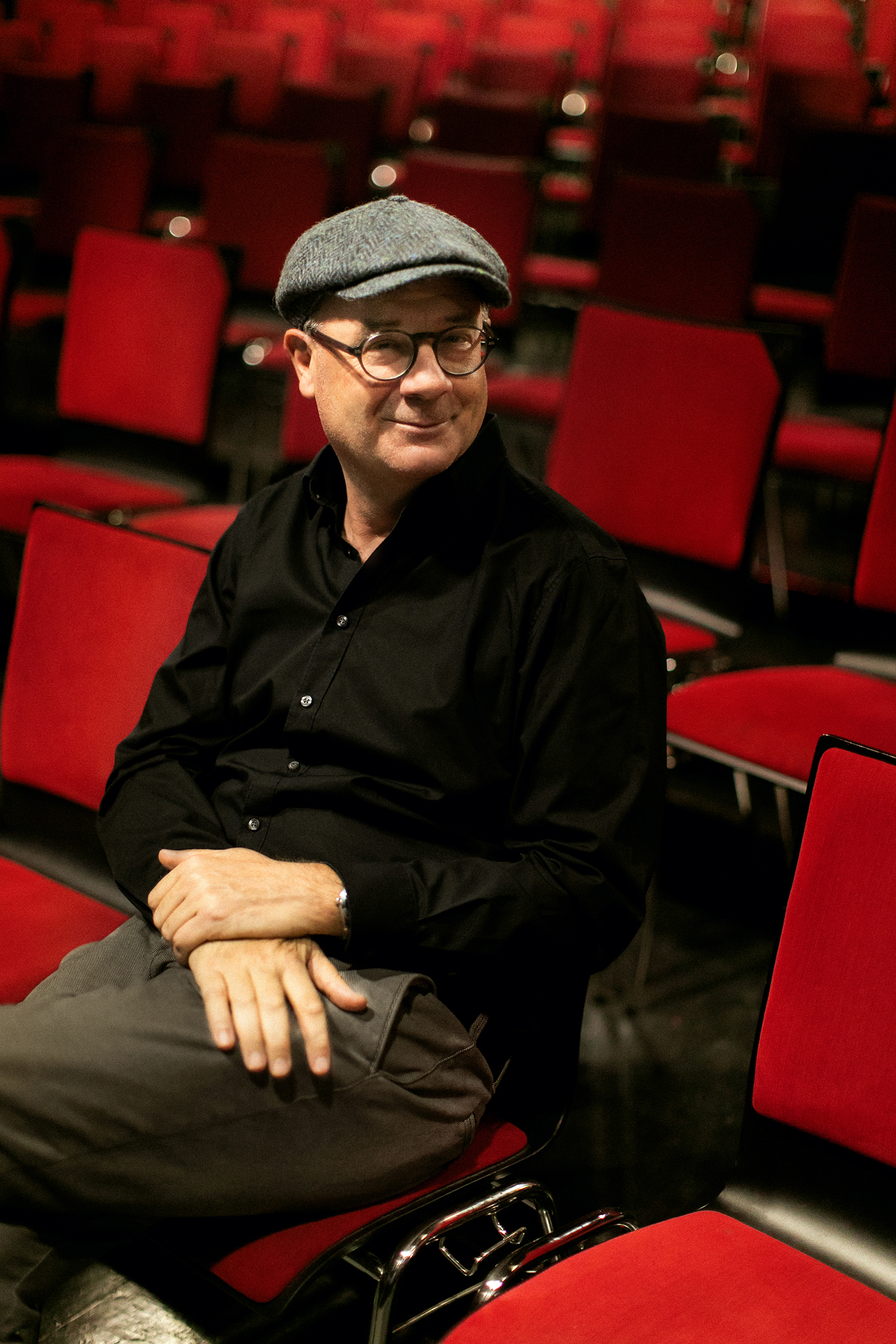 Ein Mann mit flacher Mütze, Brille und schwarzem Hemd sitzt auf einem roten Stuhl in einem leeren Hörsaal mit Reihen roter Stühle hinter ihm.