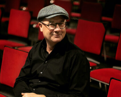 Ein Mann mit einer flachen Mütze, einer runden Brille und einem schwarzen Hemd sitzt in einem schwach beleuchteten Raum zwischen leeren roten Stuhlreihen und lächelt.