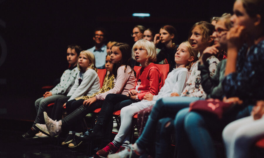 Eine Gruppe von Kindern und einige Erwachsene sitzen dicht beieinander auf Sitzen im Theaterstil und beobachten aufmerksam etwas in einem abgedunkelten Raum. Ihre Mimik reicht von neugierig bis engagiert.