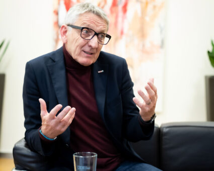 Ein Mann mit kurzen grauen Haaren und Brille sitzt auf einer schwarzen Couch und gestikuliert mit beiden Händen, während er spricht. Er trägt einen dunklen Blazer über einem weinroten Rollkragenpullover. Im Hintergrund ist ein verschwommenes abstraktes Gemälde zu sehen.