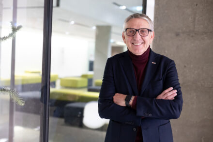 Ein lächelnder älterer Mann mit Brille, der einen dunklen Blazer und einen kastanienbraunen Rollkragenpullover trägt, steht mit verschränkten Armen in einem Haus in der Nähe einer Glaswand. Im Hintergrund sind moderne Büromöbel zu sehen.