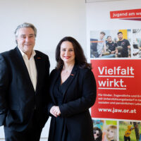 Zwei professionell gekleidete Erwachsene stehen lächelnd neben einem farbenfrohen Banner mit der Aufschrift "Vielfalt wirkt" und "jugend am werk", das für Unterstützungsdienste für Jugendliche und Erwachsene in Österreich wirbt. Auf dem Banner sind Bilder von verschiedenen Menschen zu sehen.