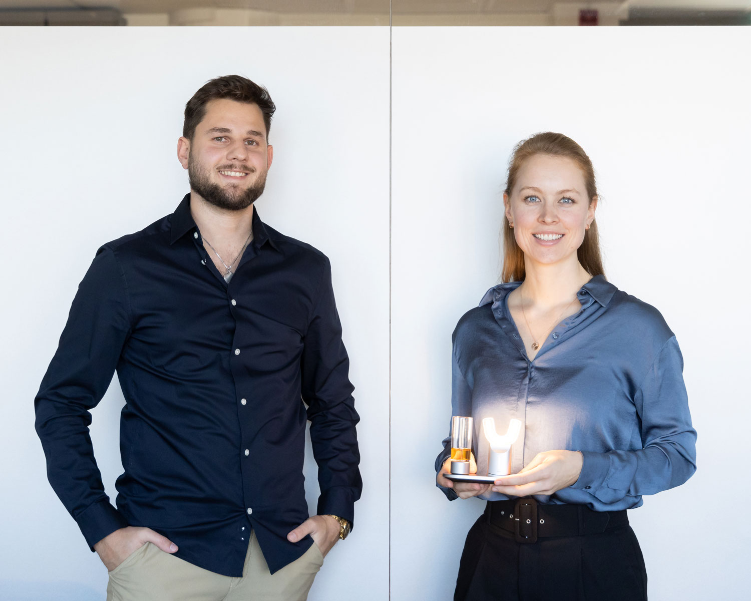 Ein Mann in einem dunklen Hemd steht neben einer Frau in einer blauen Bluse, die ein beleuchtetes Objekt in Form eines "Y" auf einem kleinen Tablett hält. Beide lächeln und stehen vor einem schlichten weißen Hintergrund.